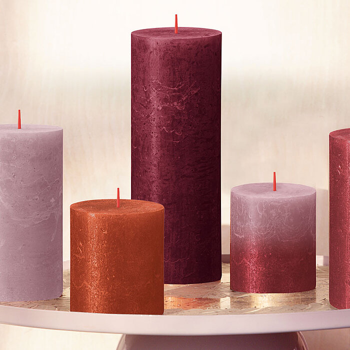 Fünf Bolsius Rustik Shimmer Kerzen, in verschiedenen Größen und Farben, stehen auf einem runden Tisch mit Spiegeloberfläche. Die Farben der Kerzen reichen von Violett über Orange bis hin zu Rot. Eine warme, einladende Atmosphäre wird erzeugt.