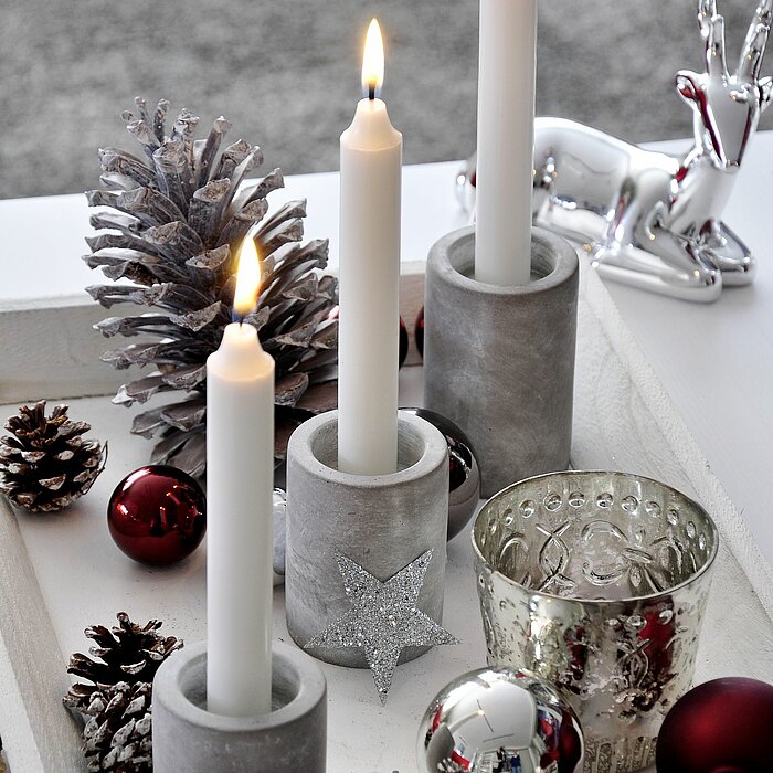 Drei brennende weiße Kerzen des Herstellers Müller in grauen, zylinderförmigen Kerzenhaltern aus Beton. Die Kerzen stehen auf einem weißen Tablett, das mit silbernen und roten Kugeln sowie Tannenzapfen dekoriert ist. Ein silbernes Rentier und ein glitzern