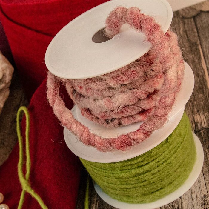 Zwei Rollen Kranzbänder vom Hersteller "Krago" auf einem Holztisch. Ein Band ist aus pink gemustertem, dicken Wollgarn, während das andere Band aus grüner, flauschiger Wolle besteht. Im Hintergrund liegt rotes und beiges Dekomaterial. Die Szene vermittelt