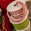 Zwei Rollen Kranzbänder vom Hersteller "Krago" auf einem Holztisch. Ein Band ist aus pink gemustertem, dicken Wollgarn, während das andere Band aus grüner, flauschiger Wolle besteht. Im Hintergrund liegt rotes und beiges Dekomaterial. Die Szene vermittelt