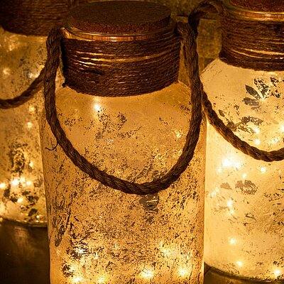 Nahaufnahme von drei LED Glasflaschen mit Korkdeckel und Sisal-Henkeln. Die Flaschen haben durch die gefrostete Oberfläche eine rustikale Oberfläche. Im Inneren leuchten LED Lichter und verbreiten ein warmes, gemütliches Licht.