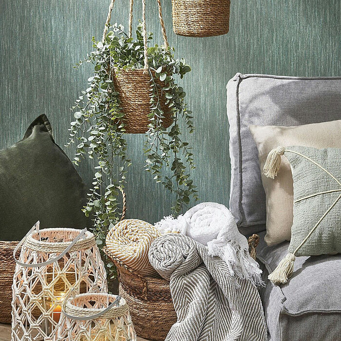 Zwei gemütlich leuchtende Laternen mit Geflechtmuster aus Seil stehen auf einem Holzboden. Daneben liegen gefaltete Decken mit Fransen in verschiedenen Mustern und ein großer grauer Sitzsack. Im Hintergrund hängen zwei Pflanzen in geflochtenen Hängetöpfen