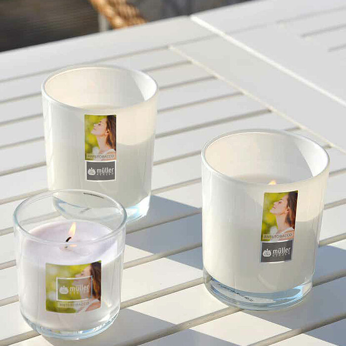 Drei brennende Kerzen des Herstellers Müller in transparenten Glasbehältern auf einem weißen Tisch. Zwei der Kerzen haben weißen Wachs, während die andere Kerze lila Wachs hat. Im Hintergrund ist eine weiße Oberfläche und ein Teil eines Stuhls zu sehen.