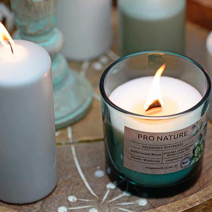 Vier brennende weiße Müller-Kerzen in Stumpenform und eine Müller-Duftkerze in einem grünlichen Glasgefäß mit der Aufschrift "Pro Nature". Die Kerzen stehen auf einem runden Holztablett. Eine der Stumpenkerzen befindet sich in einem türkisgrünen Kerzenhal
