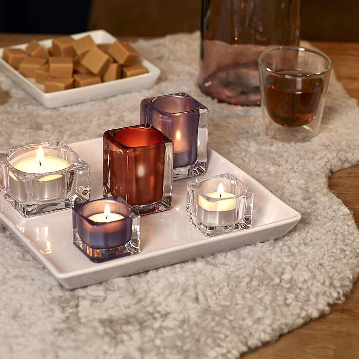 Acht brennende Teelichter in unterschiedlich gestalteten transparenten und weißen Teelichthaltern stehen auf einem weißen Tablett. Auf dem Tablett befindet sich ebenfalls eine weiße Vase mit weißen Blumen. Das Arrangement steht auf einem weißen Tischtuch.