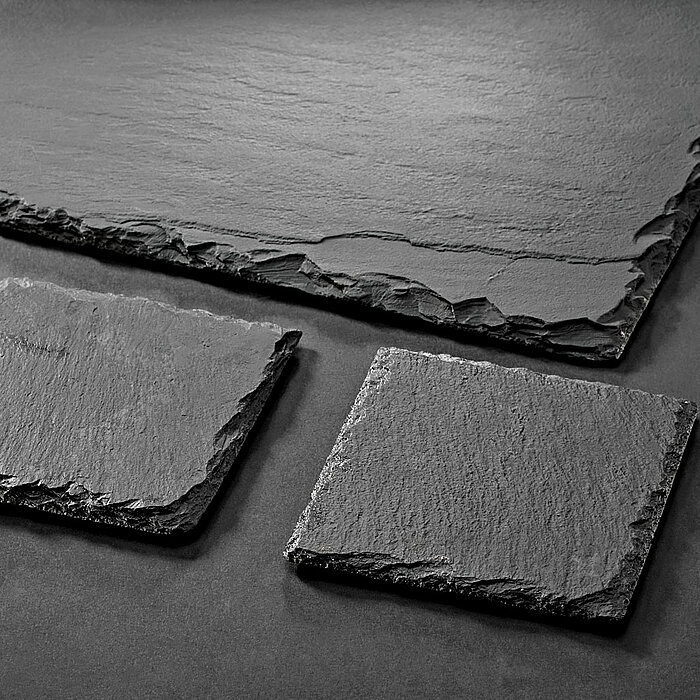 Zwei quadratische Tafeln aus schwarzem Schiefer liegen diagonal versetzt nebeneinander auf einer gleichfarbigen Oberfläche. Oberhalb der beiden kleineren Tafeln befindet sich eine größere rechteckige Schiefertafel. Die unregelmäßigen und strukturierten Rä
