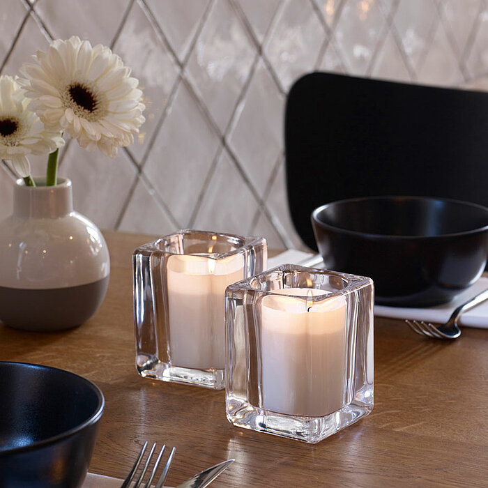 Zwei brennende rote Bolsius Kerzen in quadratischen Glasbehältern stehen nebeneinander auf einem Holztisch. Daneben befindet sich eine kleine Glasvase mit roten Blumen. Im Hintergrund sind Holz-Esstische mit weißen, geometrisch gemusterten Stühlen zu sehe