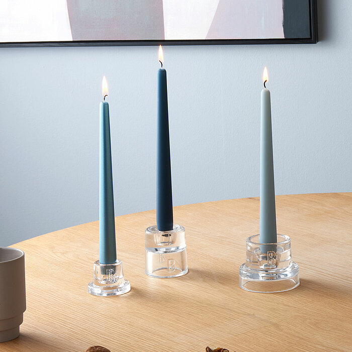 Drei brennende Bolsius Spitzkerzen in verschiedenen Blautönen stehen in klaren, modernen Kerzenhaltern auf einem runden Holztisch. Auf dem Tisch liegen zudem einige Nüsse und Nussschalen verstreut. Im Hintergrund ist ein Teil eines gerahmten Kunstwerks zu