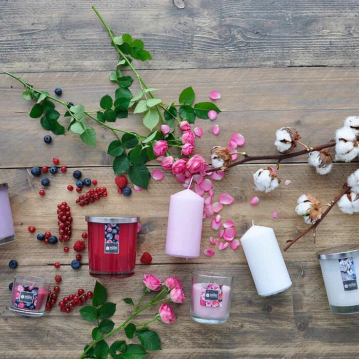Auf einem rustikalen Holztisch sind verschiedene Kerzen des Herstellers "Müller" in einer Mischung aus Glas- und Stumpenvarianten in den Farben Weiß, Rosa und Rot arrangiert. Dazu sind dekorative Elemente wie Baumwollzweige, Rosen, Beeren und Lavendel ver