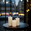 Drei weiße brennende Stumpenkerzen stehen auf einem runden Holztisch. Im Hintergrund ist eine Fensterfront zu sehen, durch die eine Stadtlandschaft mit unscharfen Lichtern und vorbeifahrenden Autos im Bokeh-Effekt erkennbar ist. Die Szenerie wirkt ruhig u