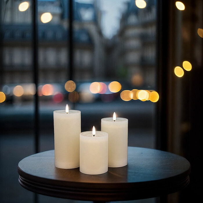 Drei weiße brennende Stumpenkerzen stehen auf einem runden Holztisch. Im Hintergrund ist eine Fensterfront zu sehen, durch die eine Stadtlandschaft mit unscharfen Lichtern und vorbeifahrenden Autos im Bokeh-Effekt erkennbar ist. Die Szenerie wirkt ruhig u