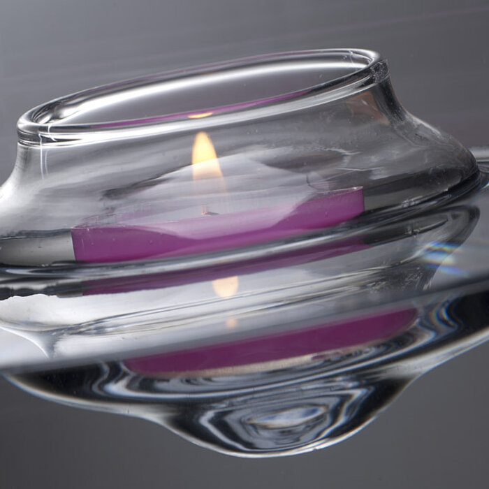 Ein einzelnes brennendes Teelicht sitzt in einem eleganten, durchsichtigen Glasbehälter. Das Teelicht strahlt in einem zarten Rosa, während das Glas klare, reflektierende Oberflächen hat, die das sanfte Licht schön verteilen. Das Teelicht und Glasbehälter