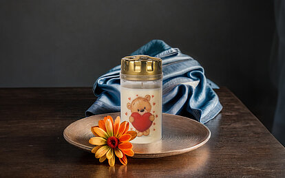 Ein Grablicht mit goldenem Deckel steht auf einem Kupferteller, zusammen mit einer orangefarbenen Blüte. Das weiße Grablicht ist mit einem Teddy verziert, der ein großes Herz in den Armen hält. Im Hintergrund ist ein blaues Satintuch zu erkennen.