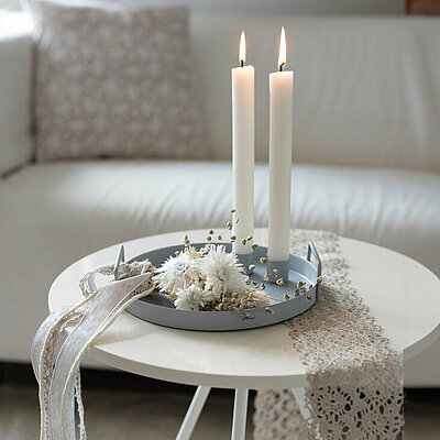 In einem gemütlichen Wohnzimmer steht eine graue gemütliche helle Couch mit einem Kissen. Im Vordergrund steht auf einem weißen Beistelltisch ein graues Metall Kerzentablett von Halbach. Auf dem Kerzentablett befinden sich zwei Magnet Kerzenhalter mit je einer brennenden weißen Haushaltskerze und einige weiße/helle Blüten und helles Stoffband als Dekoration.