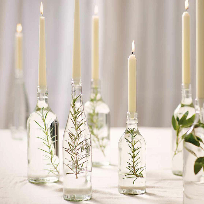 Elf brennende Bolsius Tafelkerzen in elegant arrangierten Glasflaschen auf einem festlich gedeckten Tisch. Die Tischdekoration umfasst grüne Blätter, weiße und rosa Blumen sowie zarte Glaswaren, die in einem filigranen Zusammenspiel eine romantische Atmos