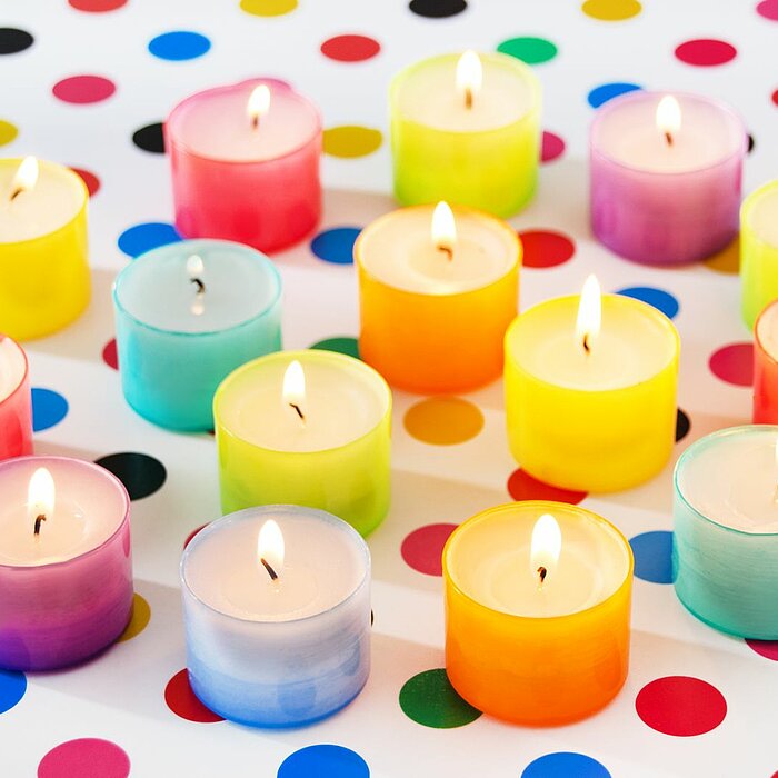 Dieses Bild zeigt fünfzehn brennende Teelichter in einer Vielzahl von Farben, darunter Gelb, Rot, Pink, Blau, Türkis, Grün, Orange und Lila. Die Teelichter sind auf einem weißen Untergrund verteilt, der mit bunten Punkten in denselben Farben wie die Kerze
