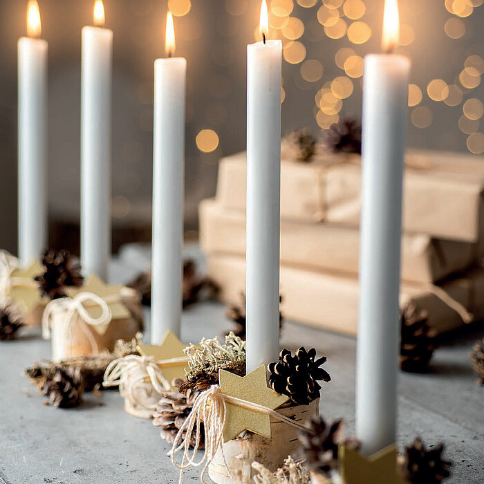 Fünf brennende, weiße Stabkerzen vom Hersteller Wenzel, die in rustikal dekorierten Haltern aus Holzscheiben und Zapfen gesteckt sind. Sie sind mit kleinen Tannenzapfen, Moos und goldenen Sternen verziert. Im Hintergrund befinden sich Geschenke in Packpap