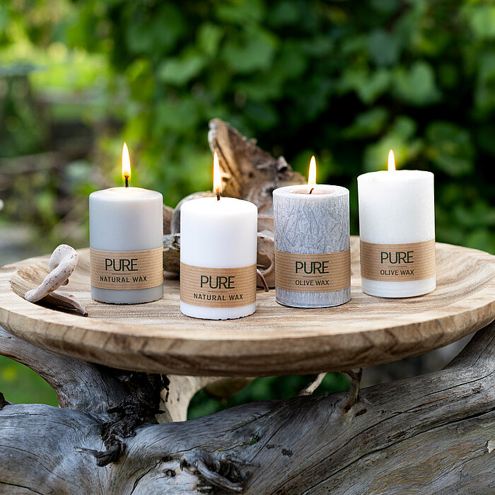 Vier brennende Kerzen vom Hersteller "Wenzel" stehen auf einer runden Holzplatte, die auf einem verwitterten Baumstamm positioniert ist. Die Kerzen sind in verschiedenen Weiß- und Grautönen gehalten und tragen die Aufschrift "PURE NATURAL WAX" und "PURE O