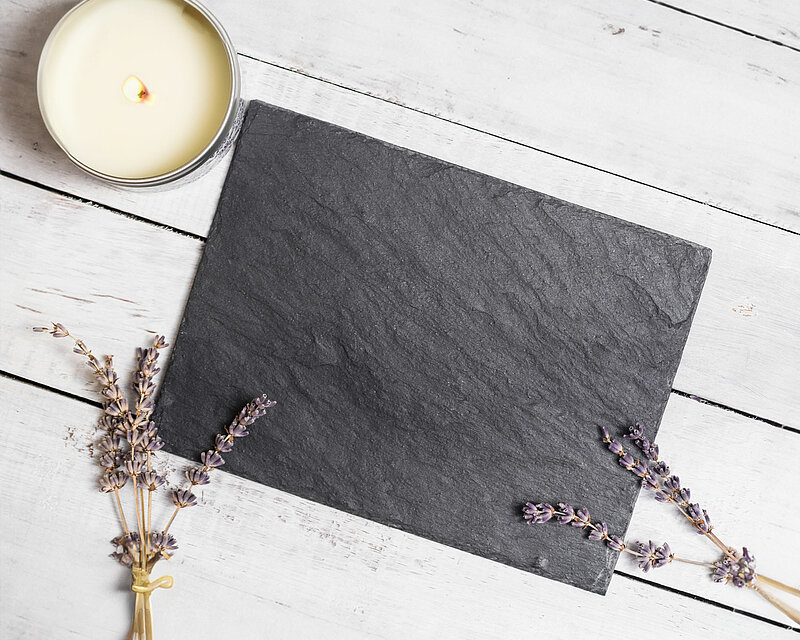 Auf einem weißen Holzboden liegt eine graue eckige Schieferplatte in anthrazit von Sandra Rich. Daneben steht eine weiße Kerze in einer Metalldose und mehrere Zweige Lavendelblüten.