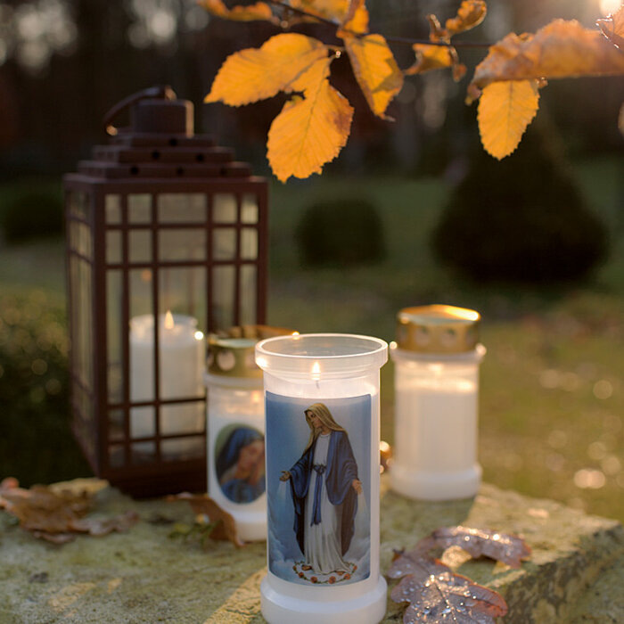 Zwei weiße Bolsius Grabkerzen: eine in einer schwarzen Laterne und die andere einfach auf einem Stein platziert. Umgeben sind sie von grünen Pflanzen und Sträuchern, die eine ruhige und friedliche Atmosphäre erzeugen.