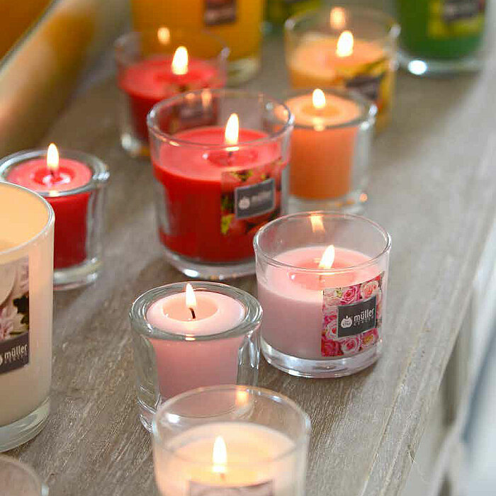 Mehrere brennende Duftkerzen des Herstellers Müller in verschiedenen Farben und Größen stehen in transparenten Gläsern auf einer Holzoberfläche. Zu sehen sind Kerzen in Rot, Weiß, Pink, Orange, Gelb und Grün. Im Hintergrund spiegelt sich eine leuchtend ge