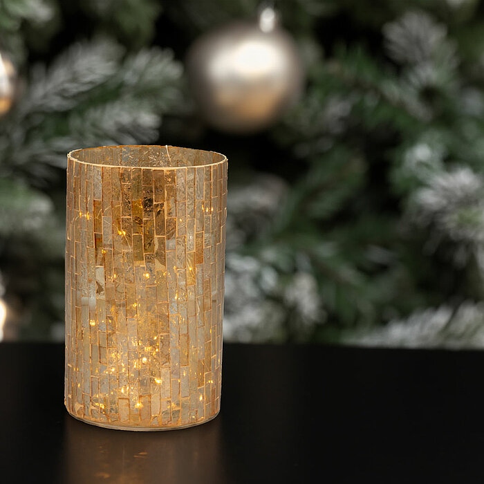 Eine goldene Kerze mit einem mosaikartigen Design aus kleinen glänzenden Fliesen brennt auf einer schwarzen Oberfläche. Im Hintergrund ist ein verschwommener Weihnachtsbaum mit silbernen und goldenen Kugeln sowie schneebedeckten Zweigen zu sehen, was eine