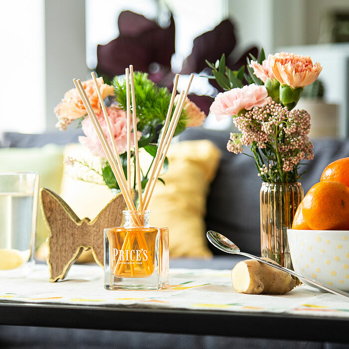 Auf einem Tisch in einem gemütlichen Wohnzimmer stehen mehrere dekorative Elemente. Im Vordergrund befindet sich eine Flasche mit Diffuserstäbchen vom Hersteller "Prices Candles". Daneben steht eine Schale gefüllt mit leuchtend orangefarbenen Mandarinen. 