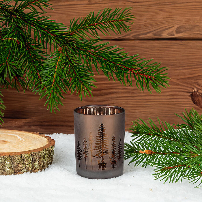 Edelman Kerzenglas mit Waldmotiv auf einem Bett aus Schnee. Das Glas steht auf einem Holzteller und ist von Tannenzweigen eingerahmt. Im Hintergrund befindet sich eine Holzwand, die dem Arrangement eine rustikale und gemütliche Stimmung verleiht.