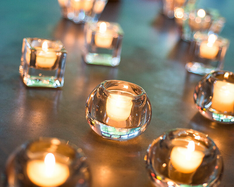 Es sind mehrere verschiedene Glas-Teelichthalter auf einer Tischplatte zu erkennen mit je einem brennenden kleinen Teelicht.