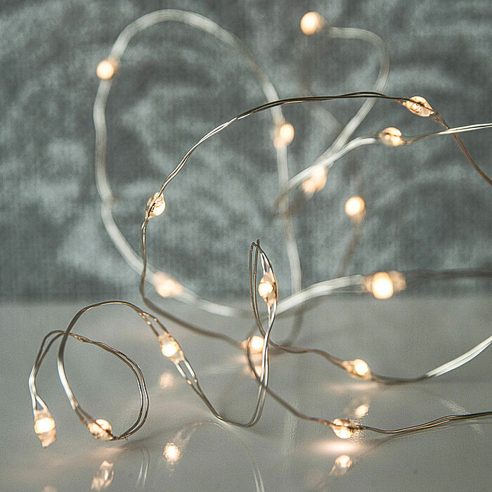 Zarte Lichterketten mit kleinen, warmweißen LED-Lampen liegen auf einer glänzenden Oberfläche und schlängeln sich kunstvoll in verschiedenen Richtungen. Der Hintergrund ist sanft unscharf und in neutralen Grautönen gehalten, was die funkelnden Lichter der