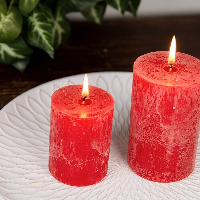 Drei brennende, rote Stumpenkerzen von DecoLite in verschiedenen Größen stehen auf einem hellen runden Tisch. Im Hintergrund sind ein braunes Sofa und cremefarbene Kissen zu sehen.