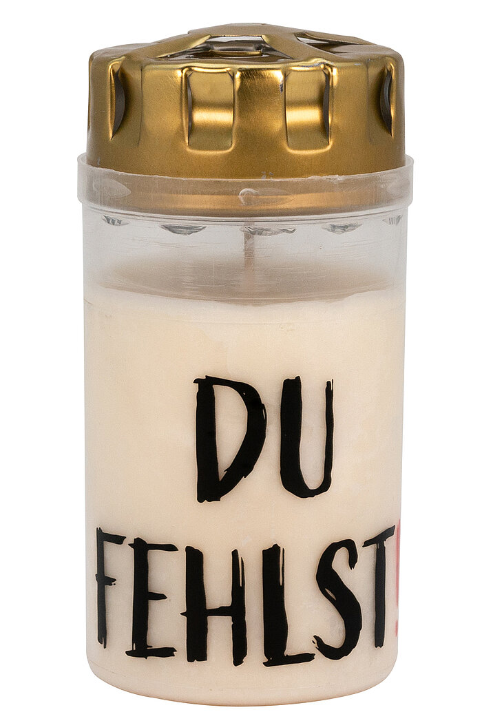 Eine weiße Gedenkkerze mit schwarzer Aufschrift "DU FEHLST" auf der Vorderseite. Die Kerze hat einen goldfarbenen Deckel mit mehreren kleinen Öffnungen.
