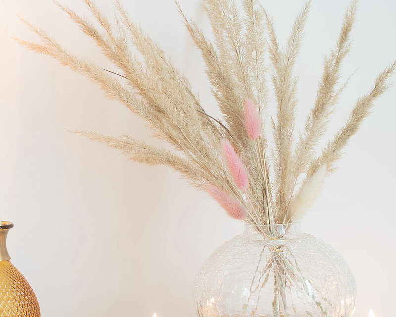 Ein farblich abgestimmtes Bild in warmen und hellen Creme und Brauntönen. Auf einem hellen Tisch steht eine sehr helle runde Holzschale. In und um die Holzschale herum verteilt sind Haselnüsse,Walnüsse, Tannenzapfen und ein paar zweige. dort steht ein brennendes Teelicht mit durchsichtigem Becher. Neben dem Teelicht steht eine bauchige durchsichtige Vase mit verschiedenen Ästen und Pampasgras. am Linken Bildrand erkennt man zur hälfte eine braune bauchige Glasvase.