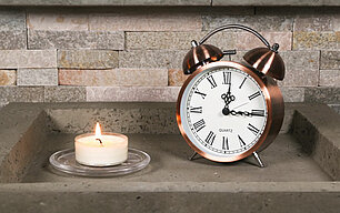 Nahaufnahme eines Teelichtes, dass in sicherer Umgebung abgebrannt wird. Daneben steht ein altmodischer Wecker, um die Brenndauer zu dokumentieren.