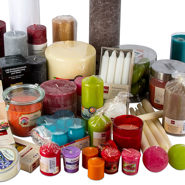 Die Aufnahme präsentiert eine beeindruckende Sammlung von Kerzen in verschiedensten Formen, Farben und Größen. Es gibt Stumpenkerzen in Grau, Rot, Gold und Blau, sowie Kugelkerzen in Lila und Rosa. Zu sehen sind auch mehrere Packungen von Stabkerzen in We