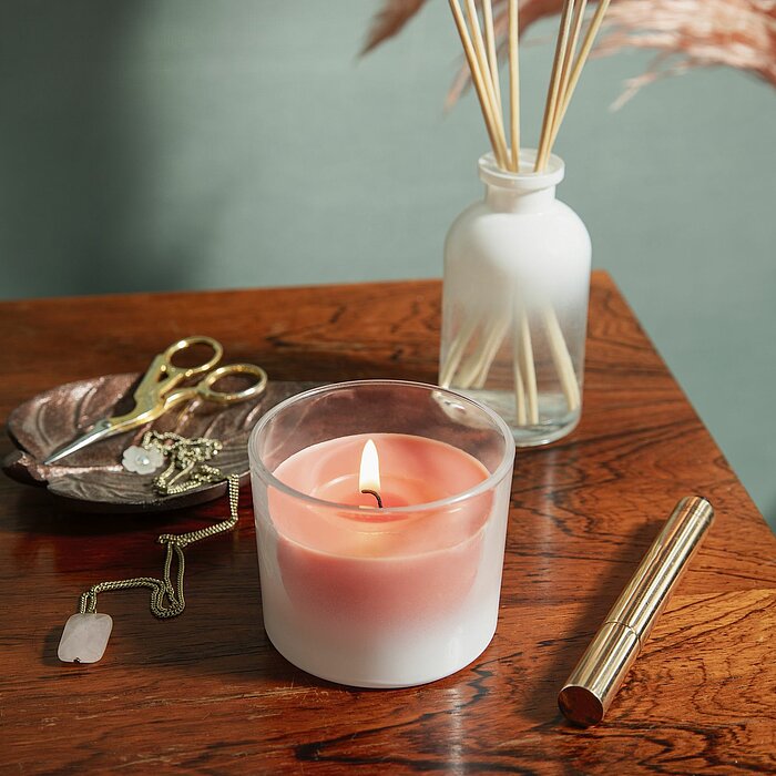 Eine Bolsius Duftkerze in einem Glas steht auf einer mintgrünen Oberfläche, umgeben von einer kleinen Blumenkomposition mit weißen Blüten und Rosen. Daneben liegt ein geöffneter Korkdeckel von Bolsius und ein Flakon mit Duftstäbchen. Halbierte Feigen und 