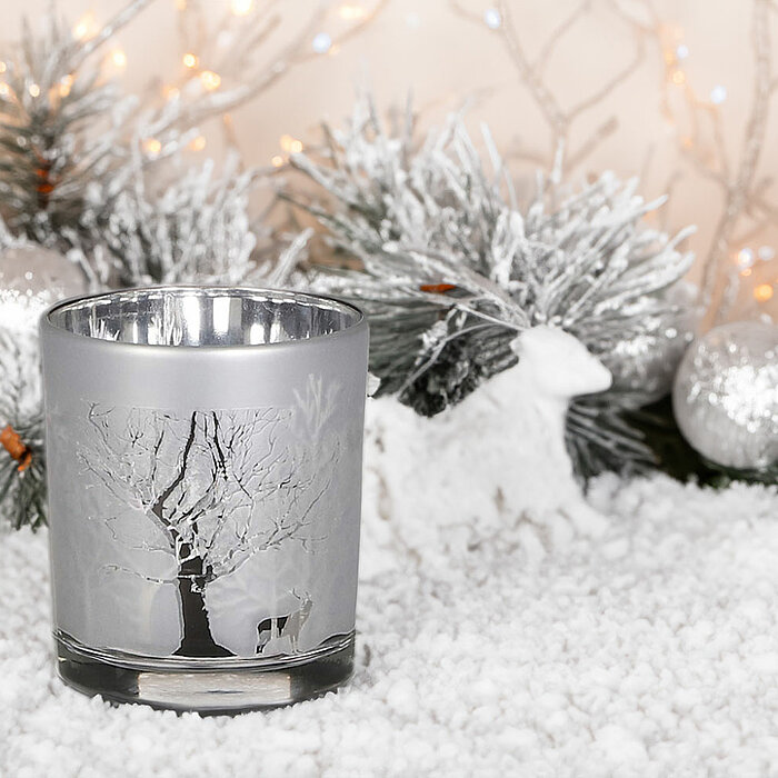 Ein Edelman Teelichthalter in Silber steht auf einer schneebedeckten Oberfläche. Der Halter ist mit einem winterlichen Baum- und Hirschmuster verziert. Im Hintergrund sind verschneite Zweige, Weihnachtskugeln und kleine, dekorative Tierfiguren zu sehen, d