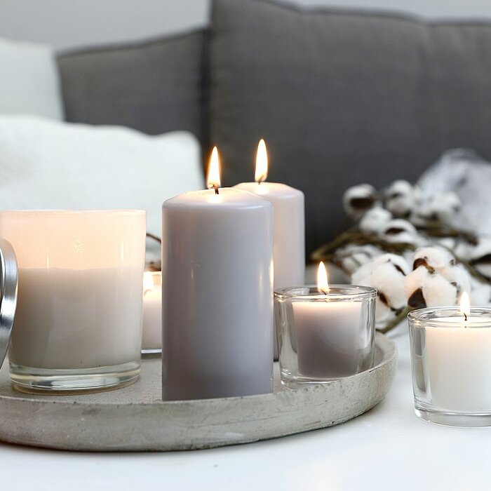 Auf dem Bild sind zwei rote Kerzengläser vom Hersteller "Müller" zu sehen, von denen eines mit einem Deckel versehen ist. Daneben brennt eine graue Stumpenkerze, ebenfalls vom Hersteller "Müller", auf einem runden Tablett. Im Hintergrund stehen ein graues