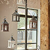Drei Laternen vom Hersteller "Edelman" stehen nebeneinander auf einem hölzernen Untergrund. Die Laternen sind in den Farben Braun, Schwarz und Weiß gestaltet und haben ein rustikales Design mit kleinen Fenstern aus Glas. Der Hintergrund zeigt ein gemütlic