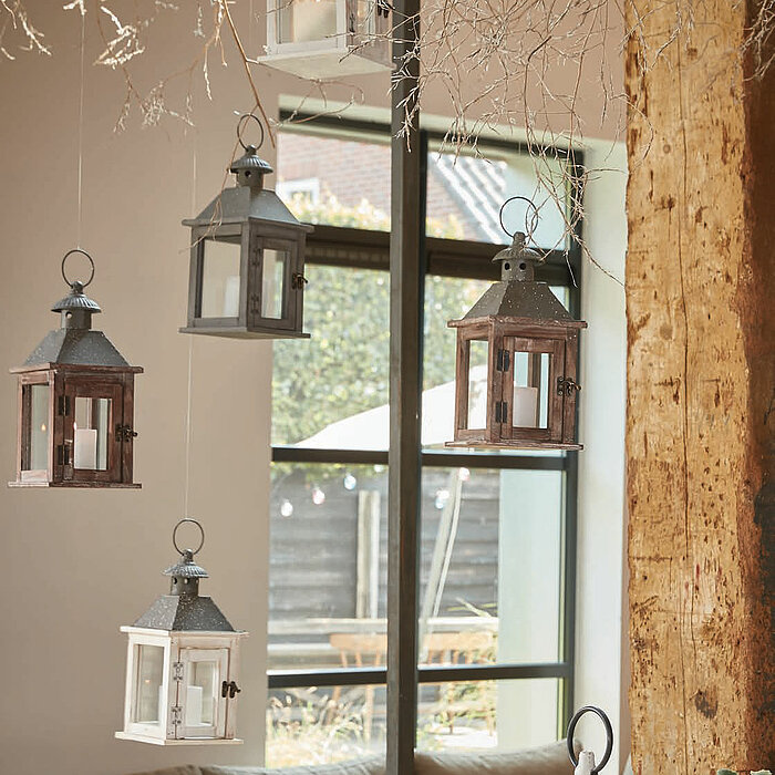 Drei Laternen vom Hersteller "Edelman" stehen nebeneinander auf einem hölzernen Untergrund. Die Laternen sind in den Farben Braun, Schwarz und Weiß gestaltet und haben ein rustikales Design mit kleinen Fenstern aus Glas. Der Hintergrund zeigt ein gemütlic