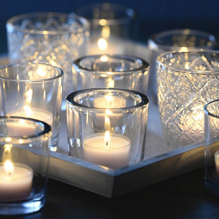 Neun brennende Teelichter vom Hersteller Müller in klaren Glashaltern, die auf einem weißen Tablett arrangiert sind. Die Glashalter variieren in ihrer Gestaltung, einige sind schlicht und andere mit Mustern verziert. Die warmen Lichter der Teelichter erze