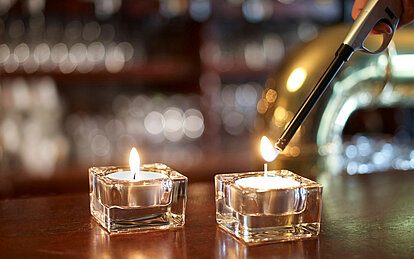 Nahaufnahme von zwei Teelichtern in einem Glashalter. Das vordere Teelicht wird gerade mit einem Stabfeuerzeug angezündet.