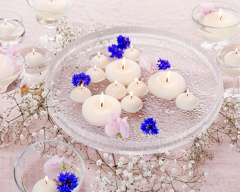 Auf einer hellen gewebten Stoffdecke sind unterschiedlich große Glasschalen platziert. Jede Schale ist mit Wasser gefüllt und enthält brennende Elfenbeinfarbene Schwimmkerzen, blaue und weiße Blüten. In der Mitte des Bildes befindet sich eine große Glasschale, ebenfalls mit Wasser und Blüten gefüllt. Auf der Wasseroberfläche schwimmen einige brennende elfenbeinfarbene Schwimmkerzen in verschiedenen Größen. Zwischen den Schalen liegen Zweige von Maiglöckchen. Die Szene strahlt ein angenehmes Kerzenlicht aus und wirkt hell, freundlich und klar.