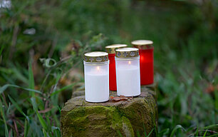 Zwei weiße und dahinter zwei rote brennende Dauerbrenner mit Deckel stehen auf einem moosigen Stein auf einer Wiese.