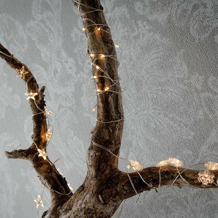 Ein Ast ist in eine glitzernde Lichterkette gehüllt, die sich um seine rauen und gebogenen Zweige windet. Die Lichterketten bestehen aus kleinen, strahlenden Lichtern, die einen warmen, gemütlichen Schein ausstrahlen. Der Hintergrund ist eine graue Wand m