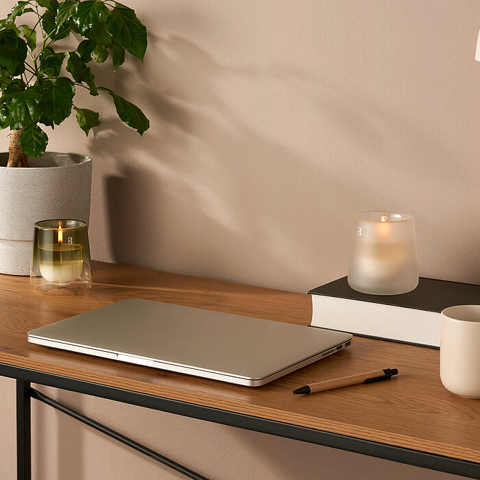 Ein modern gestalteter Schreibtisch mit einem Laptop, einem Stift und einer Tasse. Auf dem Schreibtisch steht ein Buch, auf dem eine brennende Bolsius Duftkerze in einem matten, weißen Glas platziert ist. Neben der Kerze steht eine grüne Pflanze in einem 