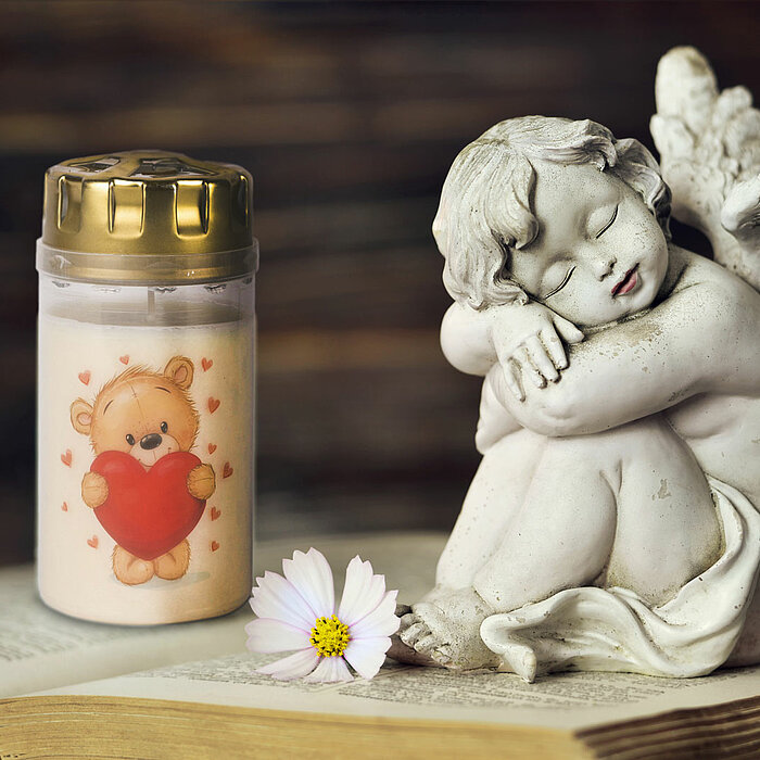 Eine weiße Gedenkkerze mit einem aufgedruckten Bärenmotiv, der ein rotes Herz hält, steht auf einem aufgeschlagenen Buch. Neben der Kerze befindet sich eine detailreiche Engelsskulptur, die die Augen geschlossen hat und die Arme um die Knie legt. Vor der 