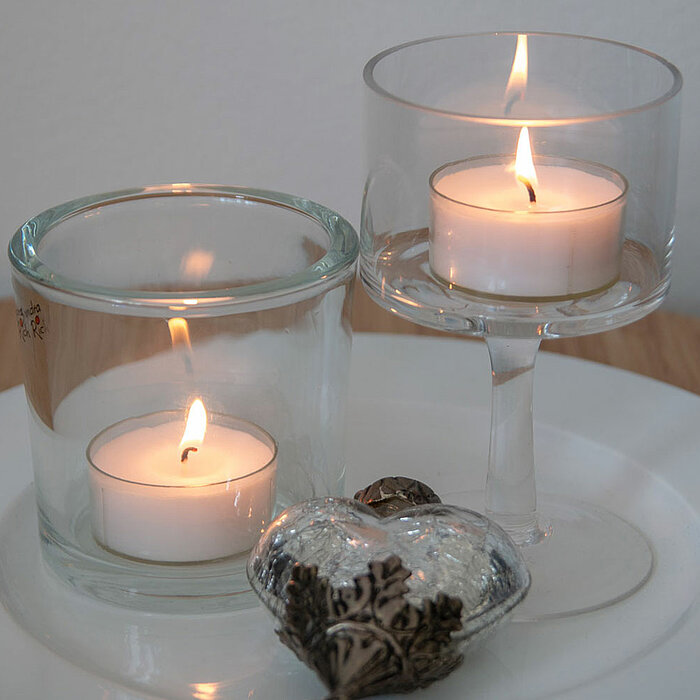Zwei brennende Teelichter in durchsichtigen Glasbehältern stehen auf einem runden, weißen Keramikteller. Ein Glasbehälter hat die Form eines Weinglases. Auf dem Teller liegt auch ein dekoratives herzförmiges, silbernes Objekt. Der Untergrund ist ein runde