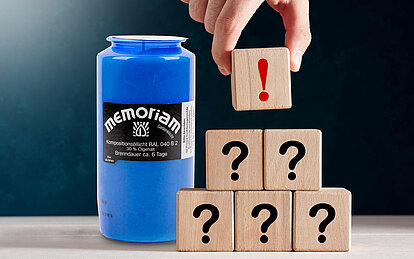 Mehrere Holzwürfel mit Fragezeichen darauf sind zu einer Pyramide gestalpelt. Oben wird gerade ein Holzwürfel mit rotem Ausrufezeichen aufgesetzt. Im Hintergrund steht eine blaue Grabkerze.