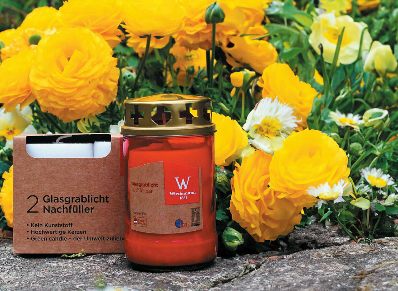 Im Vordergrund auf der linken Seite steht ein Glas-Grablicht aus rotem Glas von Wiedemann. links daneben steht eine braune Verpackung mit zwei weissen Glasgrablicht Nachfüllern von Wiedemann. Dahinter sieht man einige gelbe Ranunkeln und grüne Knospen und einige Gänseblümchen. Alles steht und liegt auf einem Steinboden.